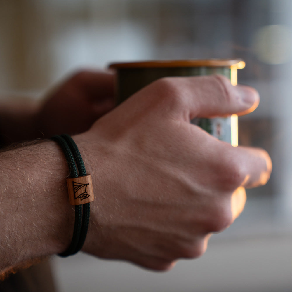Verstellbares Outdoor-Armband aus dunkelgrünem Segeltau mit Holzschieber und Camping-Gravur (Zelt im Wald) mit Schiebeverpackung.
